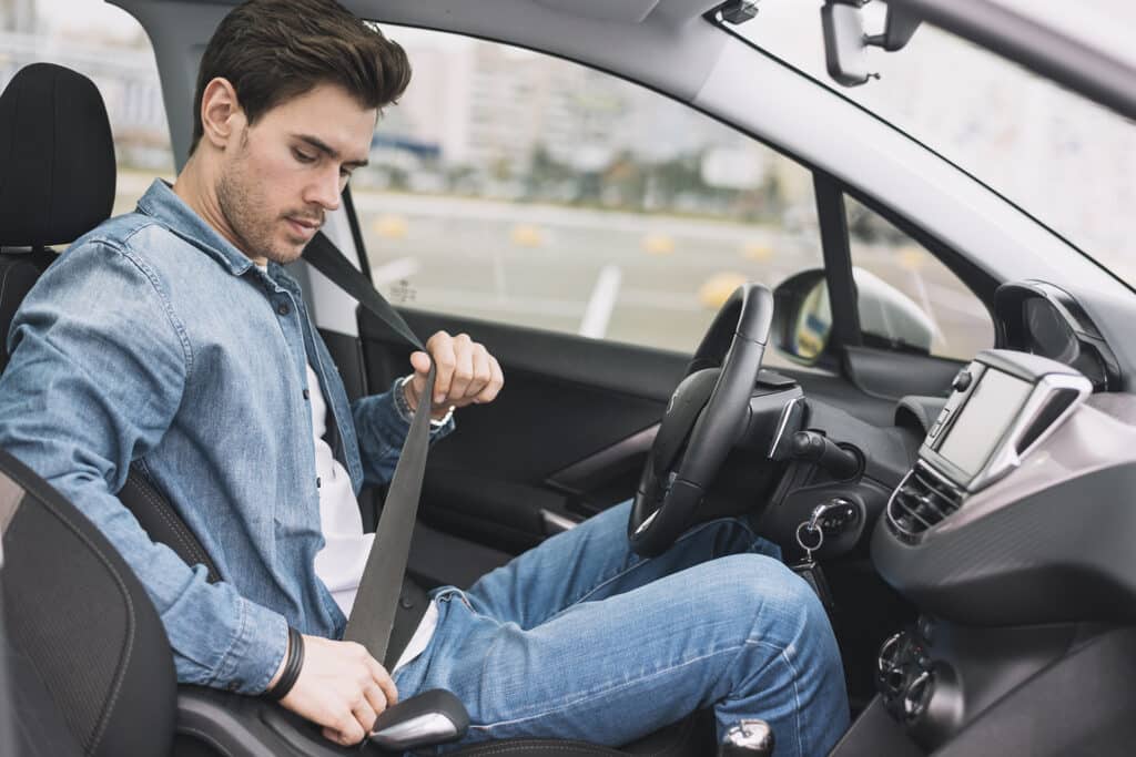 En man klädd i jeansjacka och jeans sitter i förarsätet i en bil och spänner fast sitt säkerhetsbälte. Bilens interiör är synlig, med stadsbilden suddig utanför fönstret.